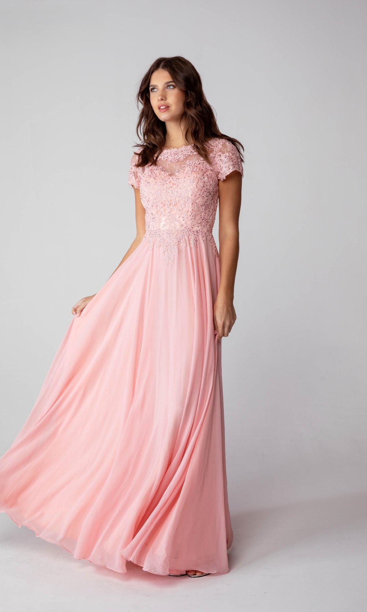 A woman poses against a plain background wearing the Eureka 4909 Short-Sleeve Long A-Line Formal Dress in light pink, featuring an embroidered lace bodice.
